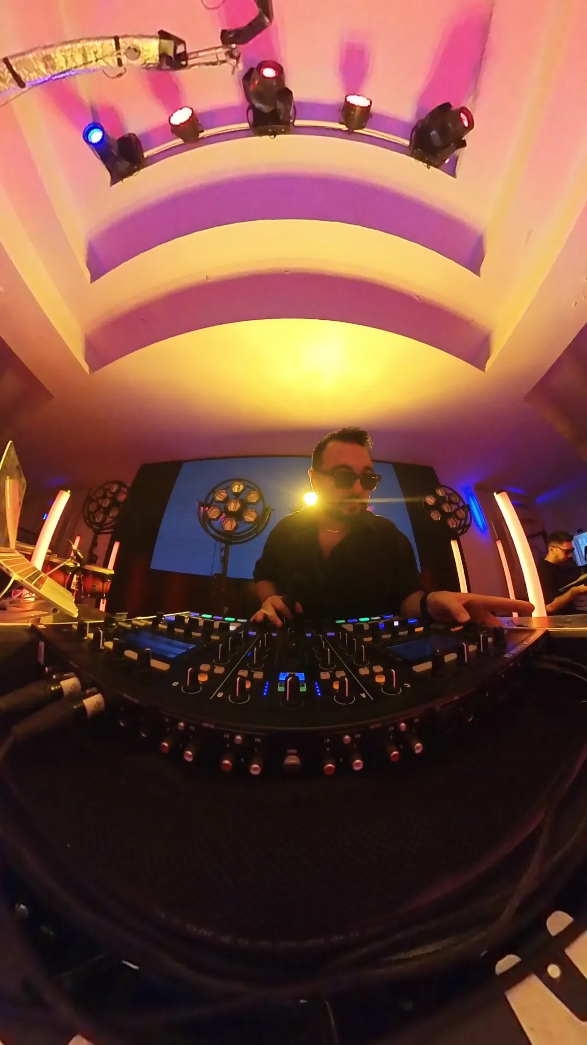 Wide booth view of DJ Mehras working the mixer under club lights